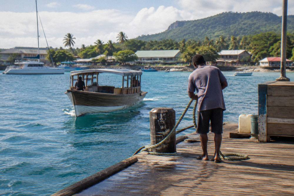 Wooden tender boat approaching a seafront jetty in Port Vila with palm trees and moored vessels
