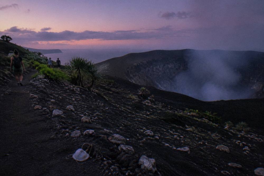 A dark volcanic slope under a wide evening sky with faint smoke drifting from the crater