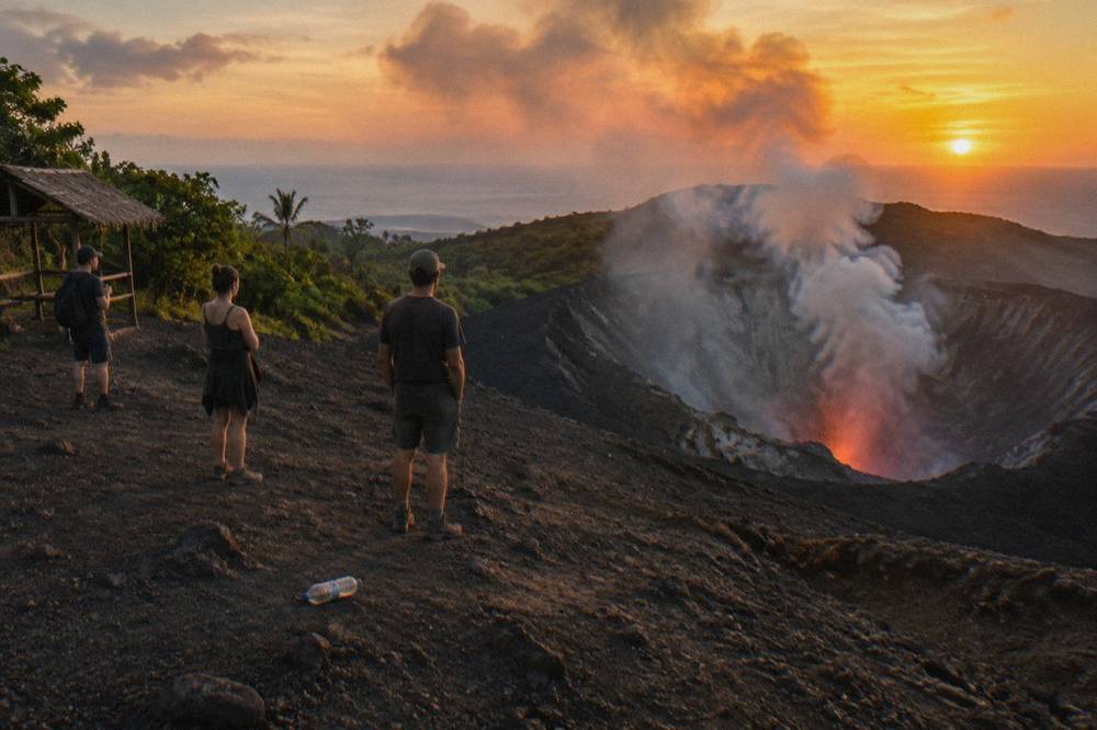 Visitors standing at a safe distance on the crater rim of Mount Yasur watching plumes rise at sunset