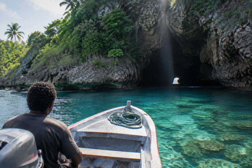 A boat approaching a dark sea cave opening in a limestone cliff above clear turquoise water