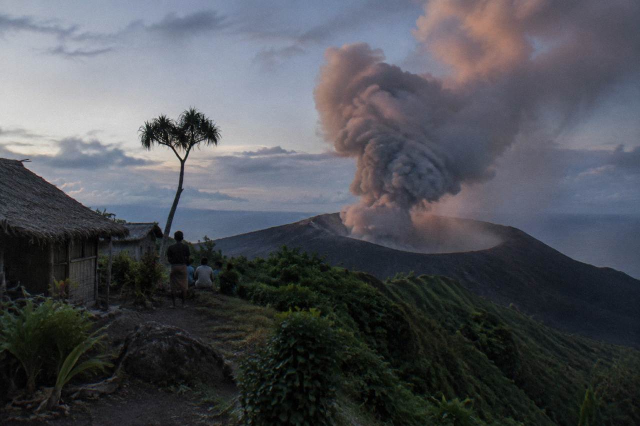 An ash plume rising from a volcanic crater above green slopes at dusk