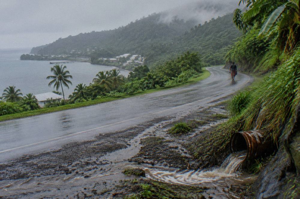 Heavy rain sweeping across a green tropical hillside with a winding coastal road below