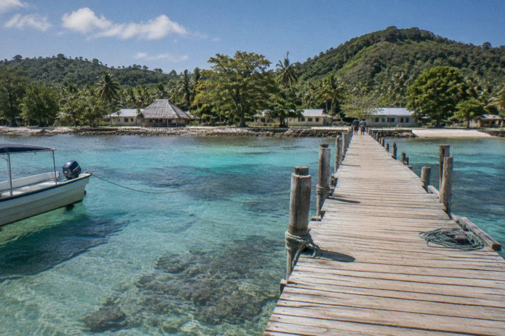 A wooden jetty extending over clear shallow reef water toward green hills on a dry sunny day