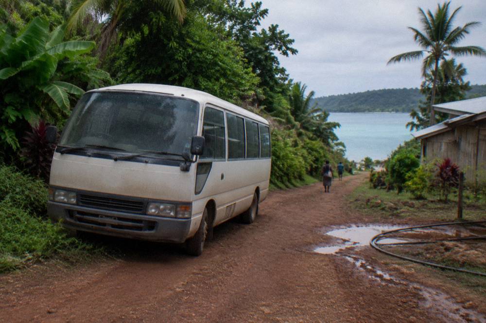 Tour van parked on a dirt track beside dense tropical vegetation near a swimming hole