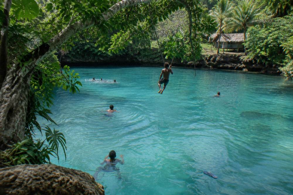 Travellers swimming in clear turquoise freshwater with overhanging trees and a rope swing