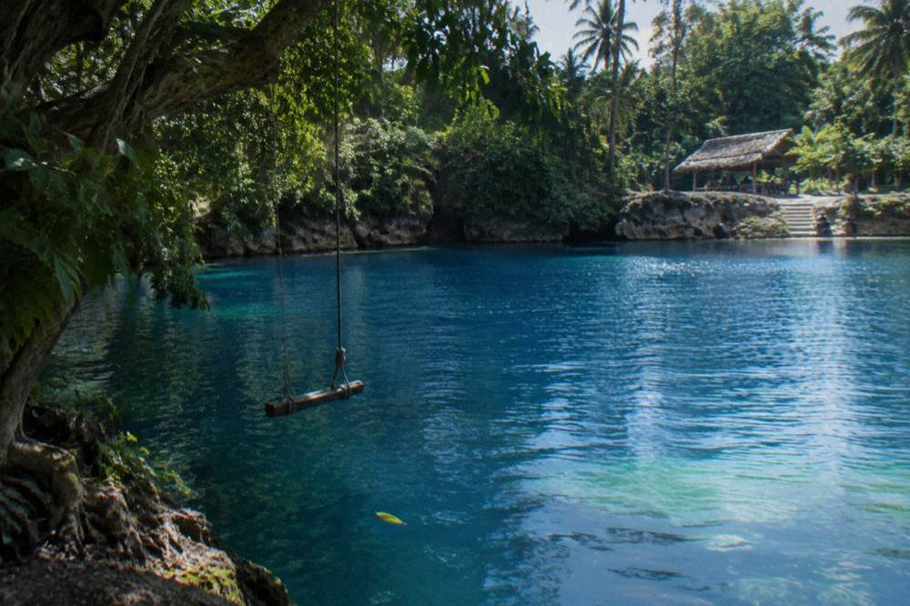 A rope swing hanging over deep blue lagoon water beneath overhanging trees