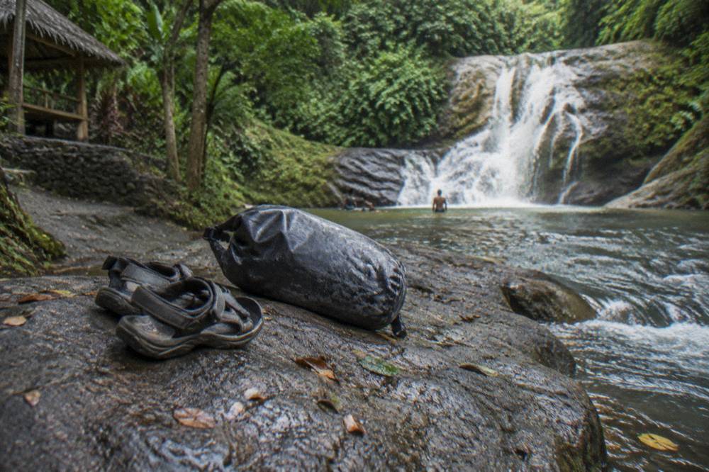 A pair of worn sport sandals and a dry bag resting on a rock beside a jungle stream