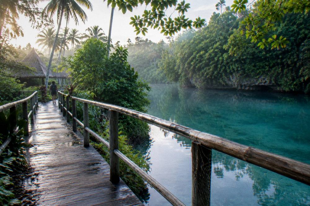 Wooden walkway leading through green jungle toward a turquoise lagoon.