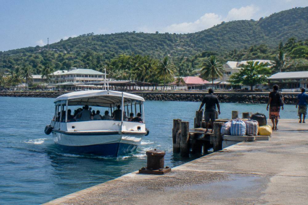 Tender boat approaching a wooden jetty beside a waterfront promenade in bright daylight