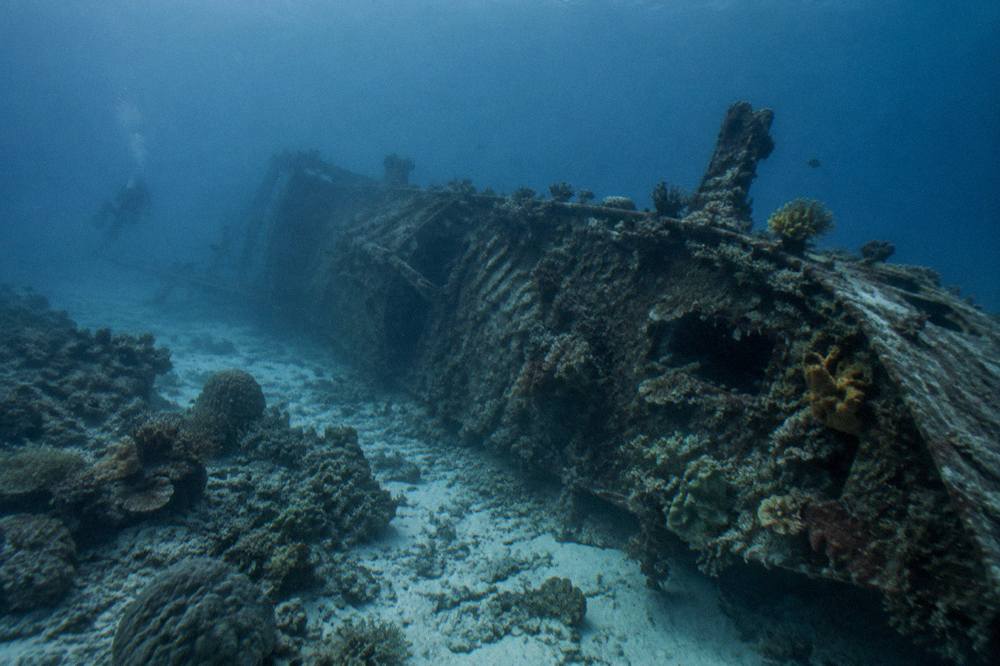 A long shipwreck hull resting on its side on an underwater slope, coral growing along the structure