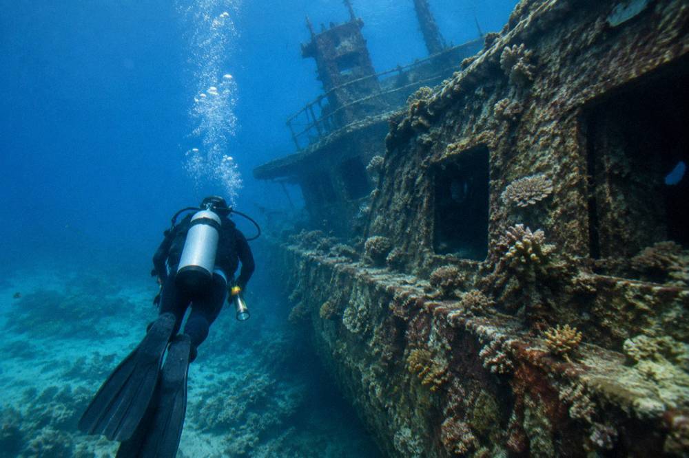 A scuba diver swimming beside a large shipwreck hull covered in coral growth