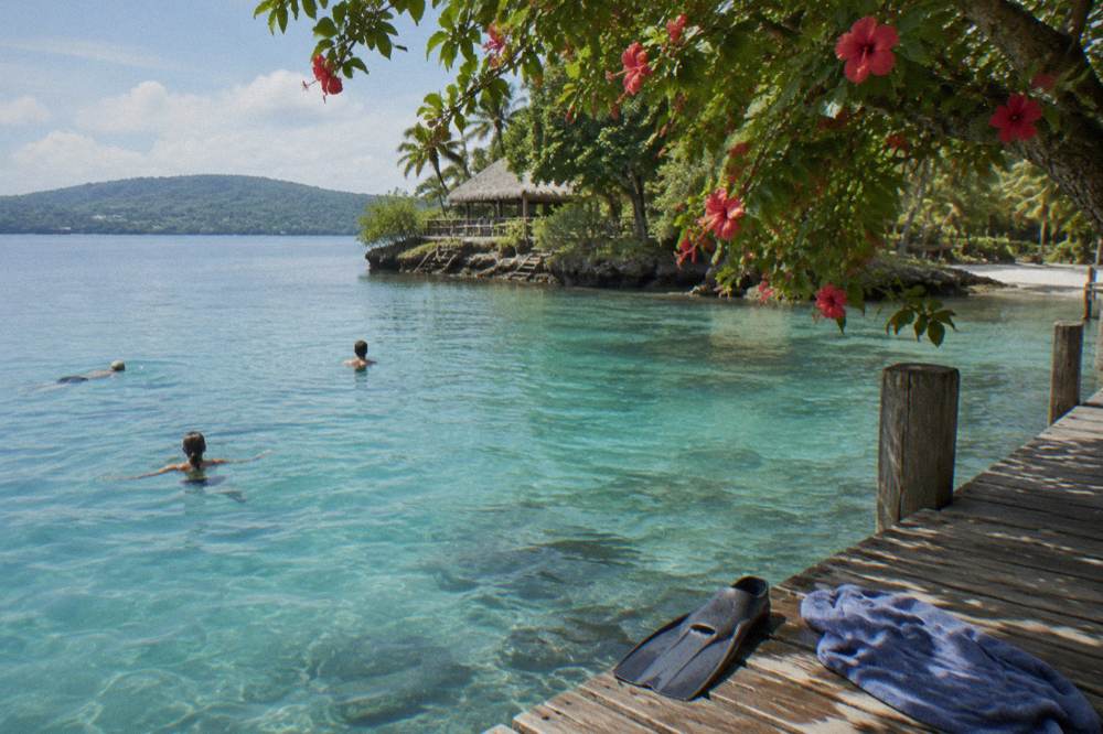 Travellers in swimwear floating in a clear blue lagoon ringed by trees and a wooden jetty