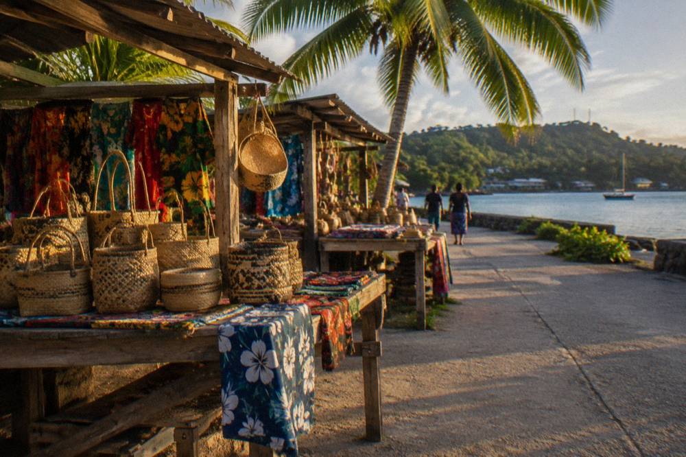 Wooden market stalls with woven baskets and fabrics set out under palm trees on a small island beach